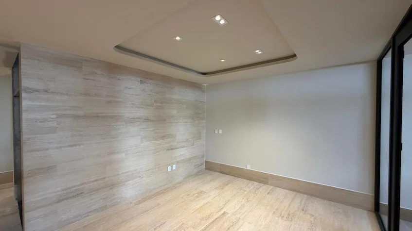 Vista interior de la sala que incluye un muro con paneles de madera clara, piso de madera, una columna con acabado de piedra texturizada y grandes ventanales con marco oscuro de Distrito Playa