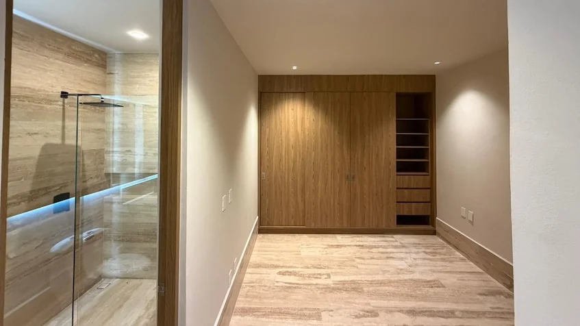Vista interior de baño y clóset que incluye un clóset amplio de madera con cajoneras abiertas y puertas abatibles, un pasillo con piso imitación madera y al fondo una zona de ducha con mampara de cristal transparente e iluminación perimetral, perteneciente al desarrollo Distrito Playa