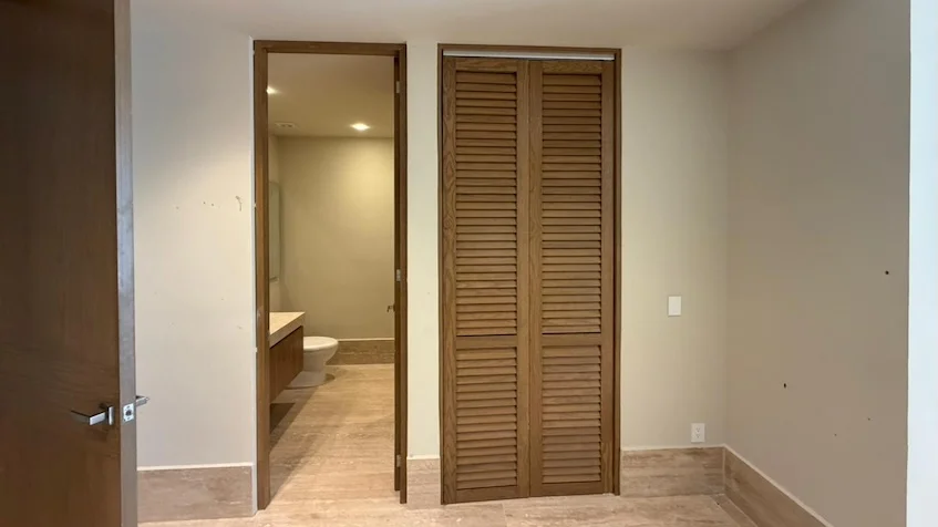 Vista de baño y clóset interior que incluye un clóset con puertas de madera de persiana, un pasillo con piso imitación madera y al fondo la vista de un baño con mueble de lavabo y taza de baño, perteneciente al desarrollo Distrito Playa