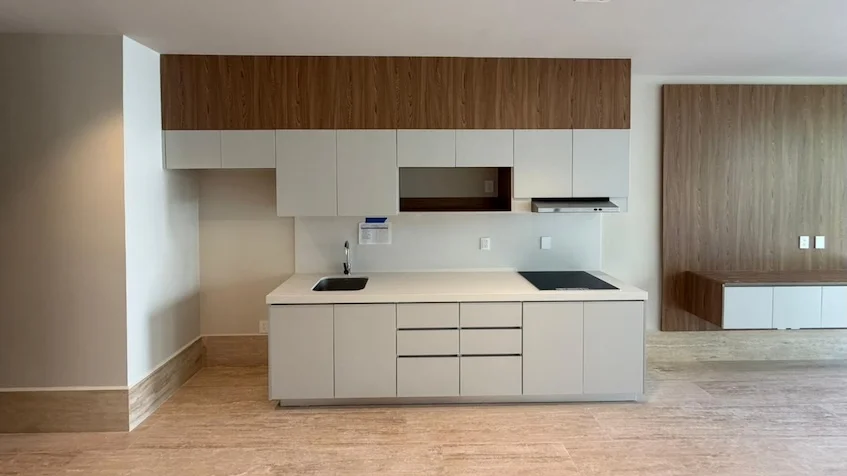 Vista interior de cocina y pasillo que incluye una barra lineal de cocina equipada con fregadero y gavetas en tonos blancos y madera, puertas de madera y piso de madera, perteneciente al desarrollo Distrito Playa