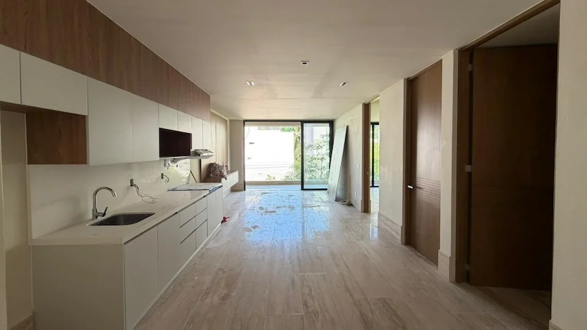 Vista interior de cocina y pasillo que incluye una barra lineal de cocina equipada con fregadero y gavetas en tonos blancos y madera, puertas de madera y piso de madera, perteneciente al desarrollo Distrito Playa