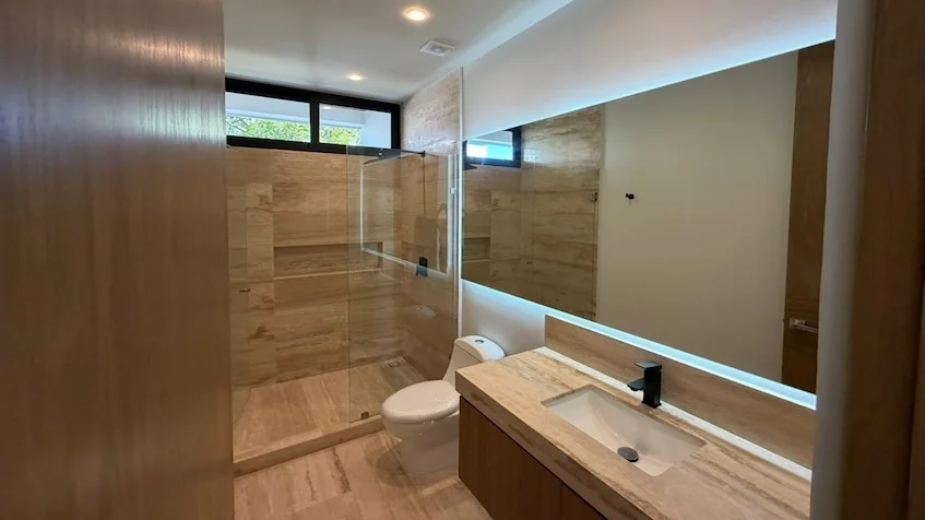 Vista de baño interior que incluye un lavabo sobre un mueble de madera, un espejo horizontal con iluminación LED perimetral, un inodoro, una zona de ducha con mampara de cristal y paredes con revestimiento de azulejos texturizados de Distrito Playa
