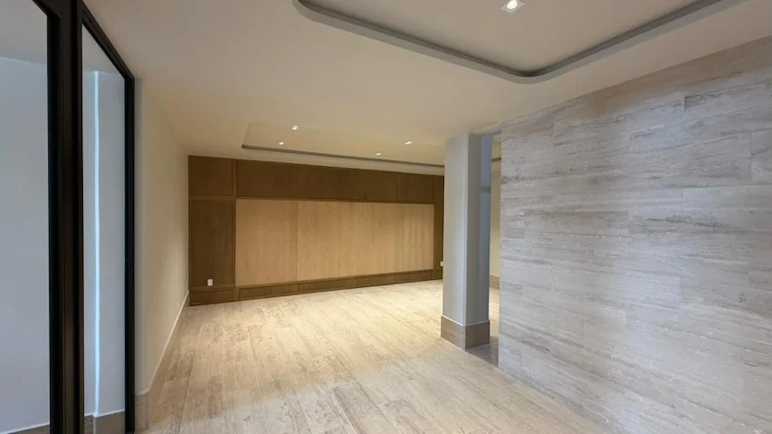 Vista interior de la sala que incluye un muro con paneles de madera clara, piso de madera, una columna con acabado de piedra texturizada y grandes ventanales con marco oscuro de Distrito Playa