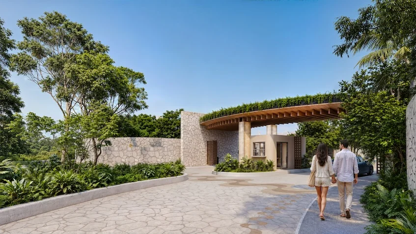 Vista del acceso principal que incluye una vialidad pavimentada, una rotonda con un árbol, un muro decorativo, una caseta de vigilancia, áreas verdes y edificios residenciales al fondo de The Origin By Gran Selva Puerto Morelos