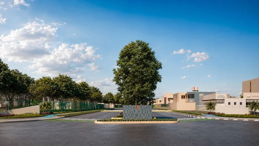 .Vista del acceso principal que incluye una vialidad pavimentada, una rotonda con un árbol, un muro decorativo, una caseta de vigilancia, áreas verdes y edificios residenciales al fondo de Vía Verde Playa del Carmen.