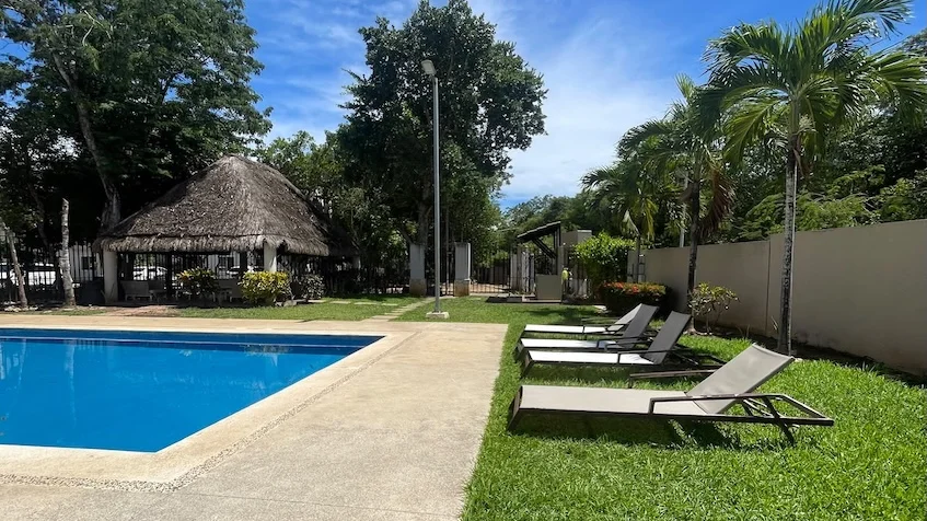 Área de alberca que incluye una piscina, camastros, áreas verdes, palmeras y una palapa al fondo de Jardines de Mayakoba Playa del Carmen.