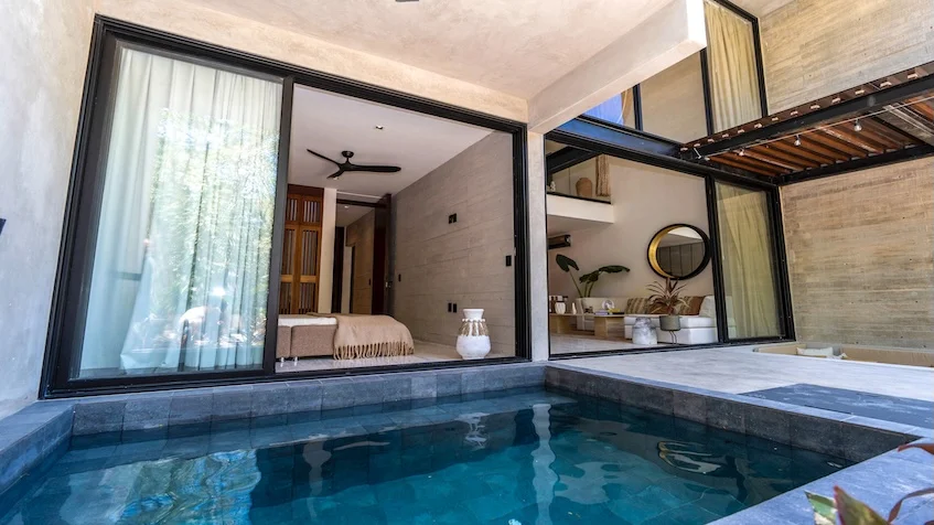 Alberca y terraza que incluye una piscina privada al aire libre, un área de descanso con piso de piedra, una pérgola de madera superior, muros de concreto aparente y grandes ventanales corredizos con vista al interior de Senzik Tulum