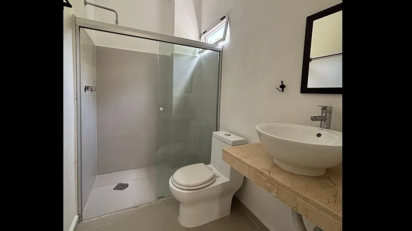 Baño que incluye una regadera con cancel de cristal, un inodoro, un lavabo sobre cubierta, un espejo y una pequeña ventana de Condominio Uxmal Playa del Carmen.