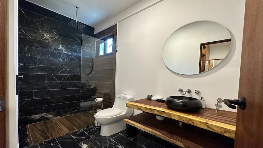 Baño interior que incluye un lavabo de piedra oscura sobre una cubierta de madera natural con borde rústico, un espejo redondo con marco de madera, un inodoro, una zona de ducha con cancel de cristal y paredes revestidas con piedra oscura de Senzik Tulum