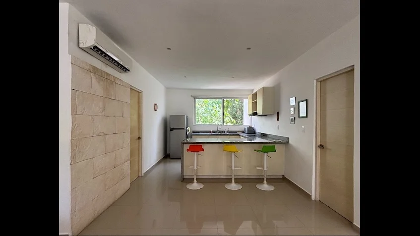 Cocina que incluye una barra con bancos de colores, un muro de piedra, un aire acondicionado, un refrigerador y una ventana de Condominio Uxmal Playa del Carmen.
