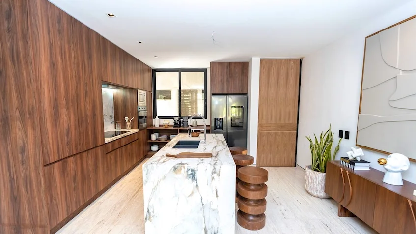 Cocina integral y barra que incluye una isla central con cubierta de mármol y base de madera, gabinetes de madera al fondo, taburetes circulares y un aparador lateral decorativo de Senzik Tulum