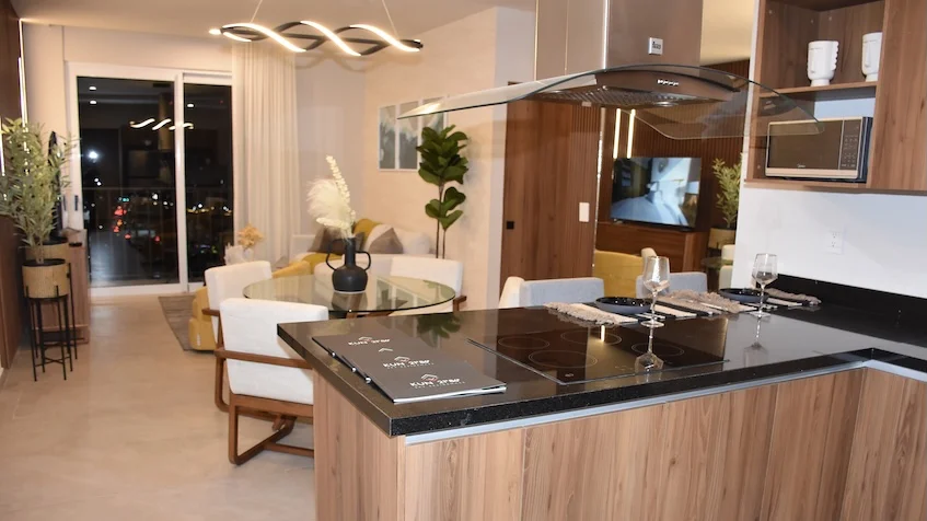 Vista interior de cocina abierta y área social que incluye una barra con cubierta de granito oscuro y parrilla integrada, campana extractora de acero inoxidable, muebles de madera, una mesa de comedor redonda con sillas tapizadas, una sala principal y grandes ventanales corredizos con vista exterior de KUN 21° 86° CancúnVista interior de cocina abierta y área social que incluye una barra con cubierta de granito oscuro y parrilla integrada, campana extractora de acero inoxidable, muebles de madera, una mesa de comedor redonda con sillas tapizadas, una sala principal y grandes ventanales corredizos con vista exterior de KUN 21° 86° Cancún