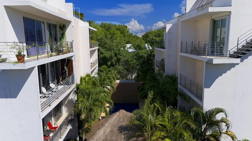 Vista exterior de edificios que incluye balcones, áreas verdes, palmeras y una palapa de Condominio Uxmal Playa del Carmen.