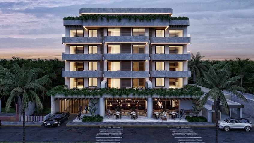 Vista frontal de edificio de condominios de cinco niveles al atardecer con balcones iluminados con vegetación colgante en cornisas planta baja con restaurante y mesas en terraza flanqueado por palmeras y al frente estacionamientos de Village Blu Puerto Morelos