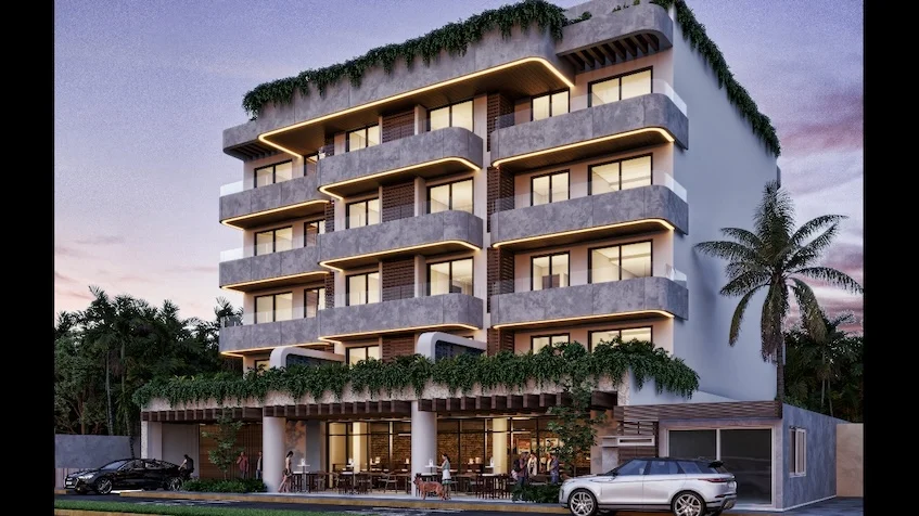 Fachada exterior de edificio residencial de varios niveles con balcones iluminados y vegetación colgante planta baja comercial con terraza y mesas al aire libre y al frente estacionamientos de Village Blu Puerto Morelos