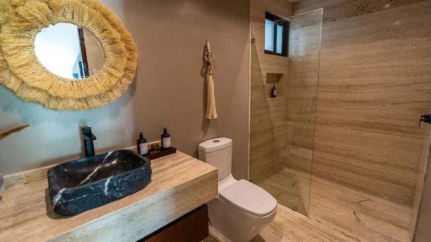Baño interior que incluye un lavabo de piedra oscura sobre una cubierta de mármol beige, un espejo redondo con marco tejido de mimbre, un inodoro, una zona de ducha con cancel de cristal y paredes con revestimiento cerámico de Senzik Tulum