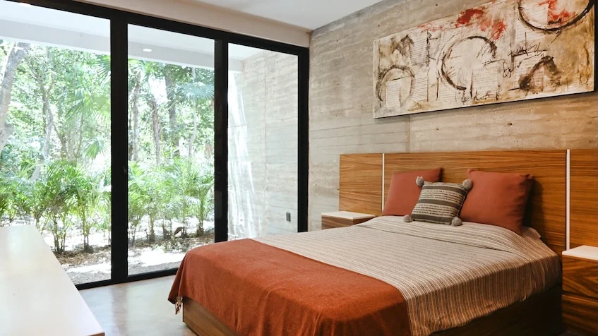 Vista de recámara con jardín que incluye una cama con ropa blanca, cabecera de madera, burós laterales, un cuadro decorativo, grandes ventanales corredizos con vista exterior a la vegetación de Senzik Tulum