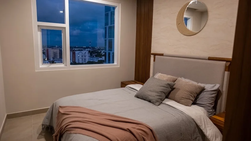 Vista de recámara que incluye una cama con ropa blanca, cojines decorativos en tonos tierra, burós laterales con lámparas, un espejo circular decorativo en la pared y grandes ventanales con vista exterior de KUN 21° 86° Cancún