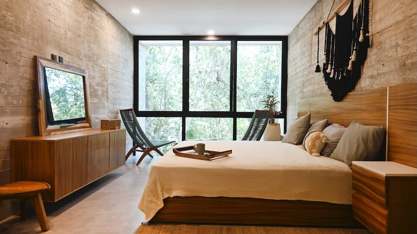Recámara secundaria que incluye una cama con ropa blanca, cabecera de madera y un adorno de macramé en la pared, burós laterales, un mueble alargado con pantalla de televisión, un sillón individual y grandes ventanales con vista a la vegetación de Senzik Tulum