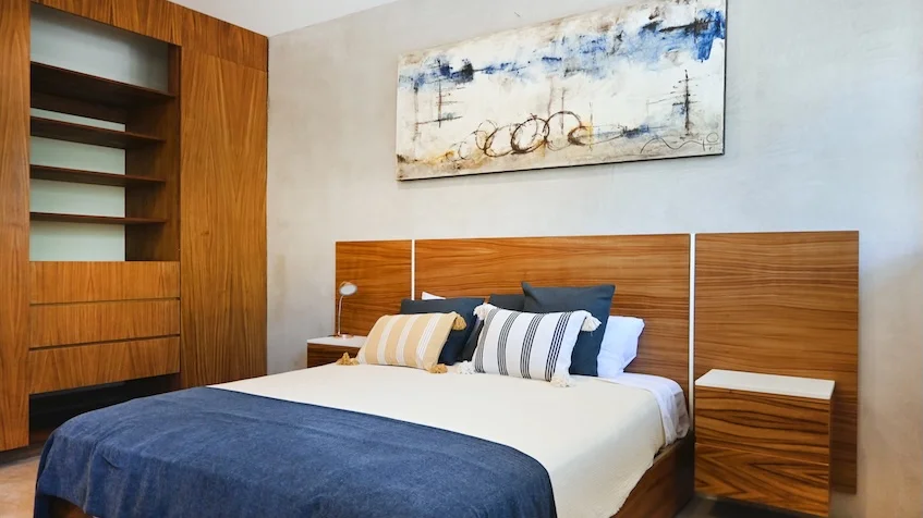 Interior de recámara que incluye una cama con ropa blanca, un clóset de madera integrado con entrepaños, burós laterales y un cuadro abstracto en la pared de Senzik Tulum