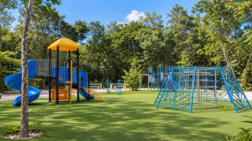 Área de juegos infantiles que incluye una estructura con resbaladilla y techo, y un domo para escalar, rodeados de vegetación de Entre Bosques Mayakobá Playa del Carmen.