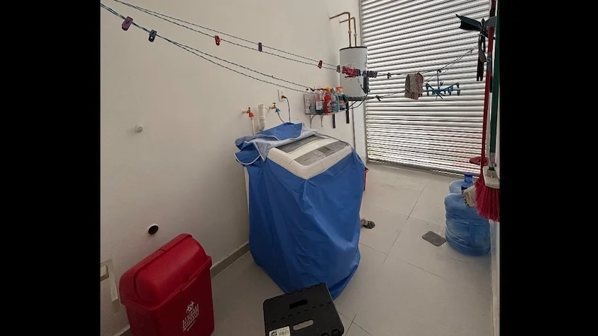 Área de lavado que incluye una lavadora con cubierta azul, un calentador de agua, un tendedero, un garrafón de agua, un contenedor rojo y una cortina metálica de Condominio Uxmal Playa del Carmen.
