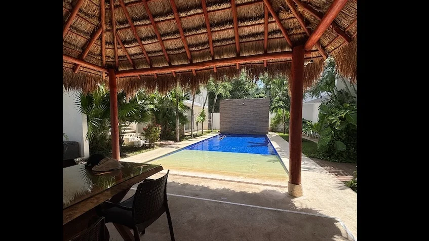 Área de alberca vista desde una palapa que incluye una mesa con sillas, una piscina, áreas verdes, palmeras y un muro de piedra de Condominio Uxmal Playa del Carmen.