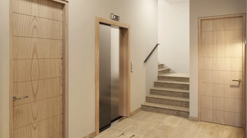 Pasillo interior de estilo moderno con piso de loseta clara ascensor central con puertas metálicas y marco de madera puertas de madera clara a ambos lados y un tramo de escaleras al fondo de Village Blu Puerto Morelos