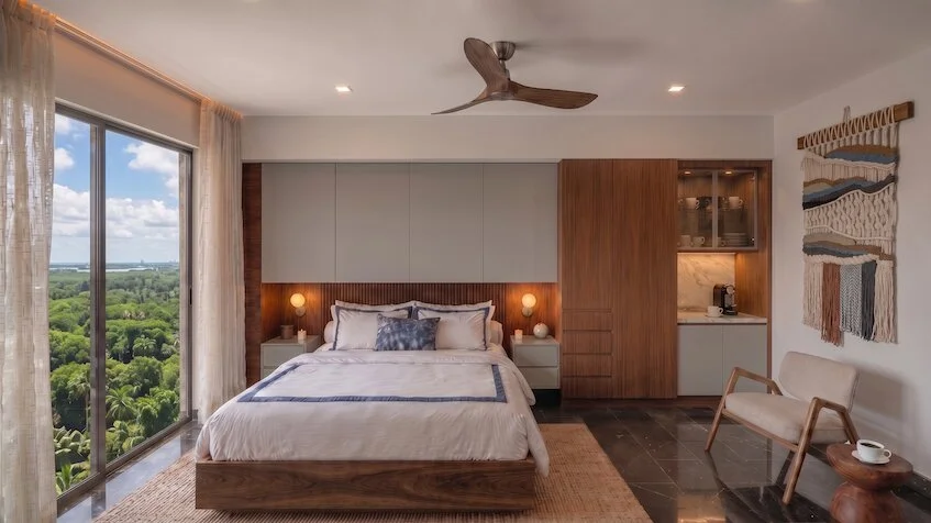Vista de recámara lock-off que incluye una cama con ropa blanca, un área de cocineta con barra y minibar, un sillón individual, grandes ventanales con vista al mar y un ventilador de techo de Sensum Puerto Morelos