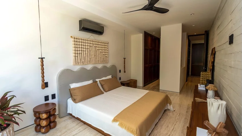 Recámara principal que incluye una cama con ropa blanca, una cabecera tapizada con un adorno de macramé en la pared, mesas de noche de madera, un ventilador de techo, un mueble de madera alargado y muros de piedra aparente de Senzik Tulum