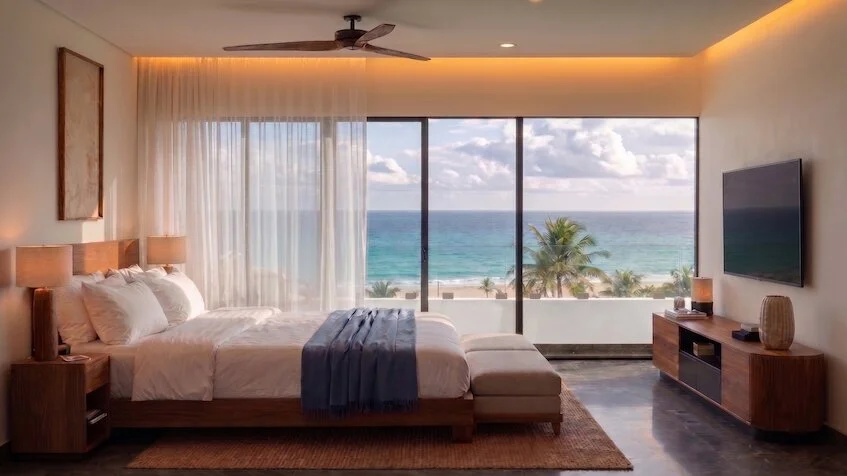 Vista de recámara que incluye una cama con ropa blanca, un mueble de televisión con pantalla, grandes ventanales y una vista panorámica al mar de Sensum Puerto Morelos