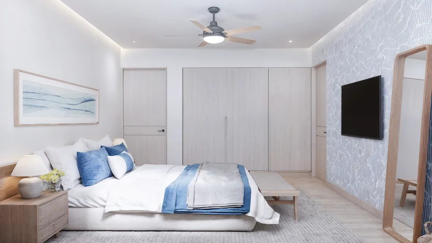 Diseño interior de la recámara principal que incluye una cama, un buró con lámpara, una banca, una televisión, un espejo, clósets y un ventilador de techo de Isola BLU Isla Mujeres.