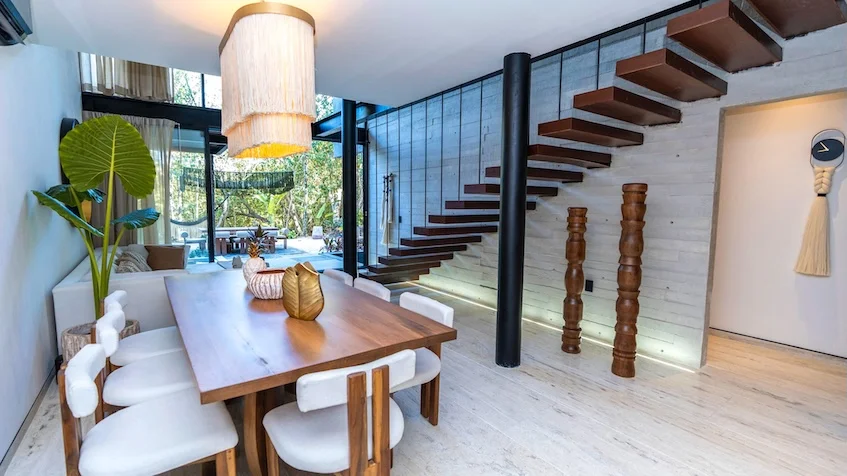 Sala y comedor que incluye una mesa de comedor de madera con sillas tapizadas, una lámpara colgante cilíndrica de mimbre, una escalera flotante de madera con barandal metálico y una sala al fondo con grandes ventanales de Senzik Tulum