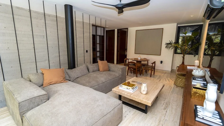 Sala que incluye un sofá gris claro con cojines ocre, una mesa de centro de madera sobre piso de mármol, un aparador lateral con elementos decorativos de Senzik Tulum