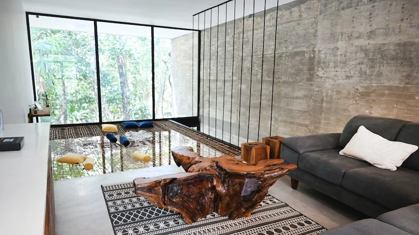 Sala de doble nivel que incluye un sofá oscuro, una mesa de centro de madera natural sobre un tapete geométrico, muros de concreto aparente, grandes ventanales con vista a la vegetación y barandales colgantes de Senzik Tulum