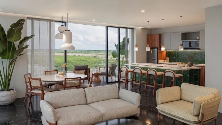 Sala y comedor que incluye dos sofás claros, una mesa de centro de madera, una mesa de comedor redonda con sillas, una cocina integral con isla central verde y grandes ventanales con vista exterior de Sensum Puerto Morelos