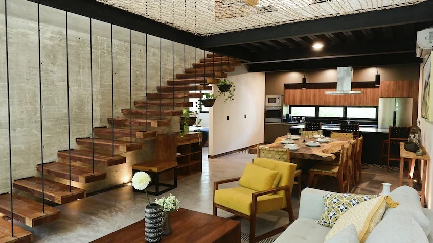 Sala y comedor que incluye un sofá claro con cojines, un sillón individual amarillo, una mesa de centro de madera, una escalera flotante con barandal de tirantes metálicos, una mesa de comedor de madera con sillas y una cocina integral equipada al fondo de Senzik Tulum