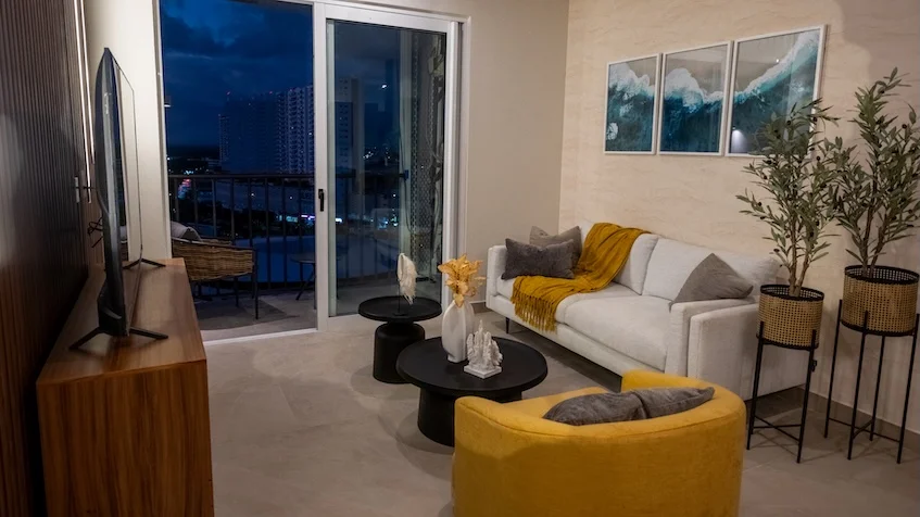 Sala de televisión que incluye un mueble de madera para pantalla, una mesa de centro circular, un sofá claro con cojines, un sillón individual amarillo, plantas decorativas en macetas, cuadros abstractos en la pared y grandes ventanales corredizos con vista exterior de KUN 21° 86° Cancún
