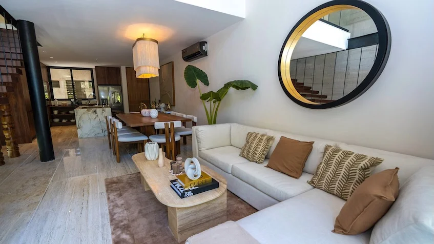 Sala que incluye un sofá seccional en tono claro con cojines decorativos, una mesa de centro de madera, un espejo circular con marco negro y dorado, un comedor de madera al fondo con sillas tapizadas y una lámpara colgante de Senzik Tulum