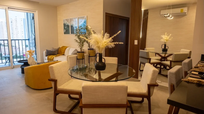 Sala y comedor que incluye una mesa de comedor redonda con sillas de madera tapizadas, una sala principal con sillón amarillo y cojines decorativos, un aparador con arreglos florales, grandes ventanales con salida al balcón y vista exterior de KUN 21° 86° Cancún