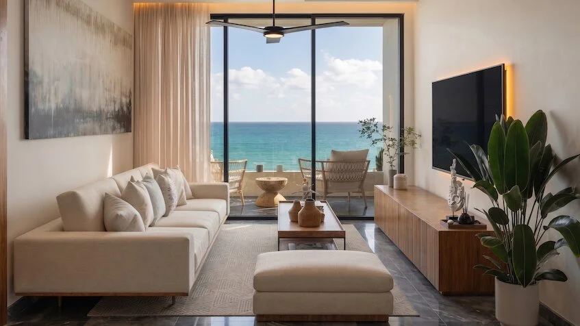 Sala que incluye un sofá beige, una mesa de centro de madera, un mueble de televisión con una planta decorativa, grandes ventanales y una vista panorámica al mar de Sensum Puerto Morelos