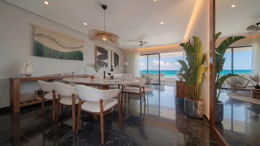 Vista de sala-comedor que incluye una mesa redonda con sillas tapizadas, un aparador de madera con elementos decorativos, grandes ventanales con vista al mar y plantas tropicales de Sensum Puerto Morelos